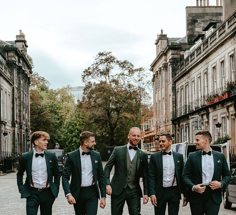 Groom and groomsman walking together in matching dark green wedding suit with black bow ties and shoes 