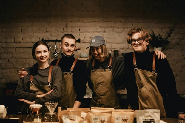 A group of four baristas smile and pose together behind their rustic-style coffee shop counter.