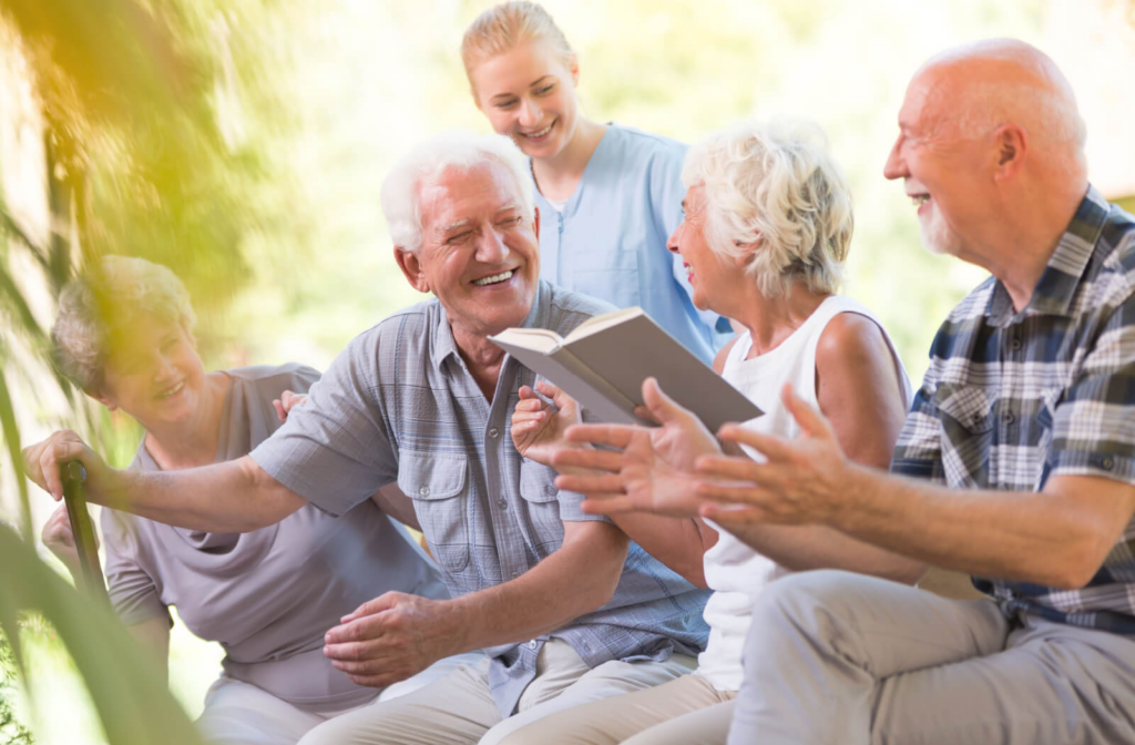 A senior man with a cane smiles and laughs with a nurse and other seniors.