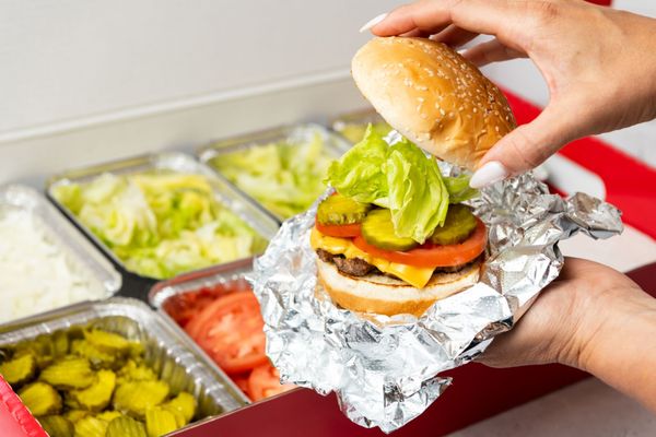 Photo of Five Guys - New York, NY, US. A close-up photo of a person putting the top bun of their cheeseburger in front of a Five Guys catering box of toppings.