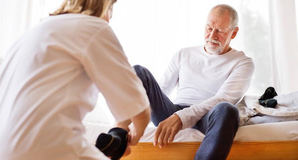 A caregiver is assisting an elderly man, who is sitting on a bed, with putting on socks, suggesting support and care for mobility or diabetic foot care.