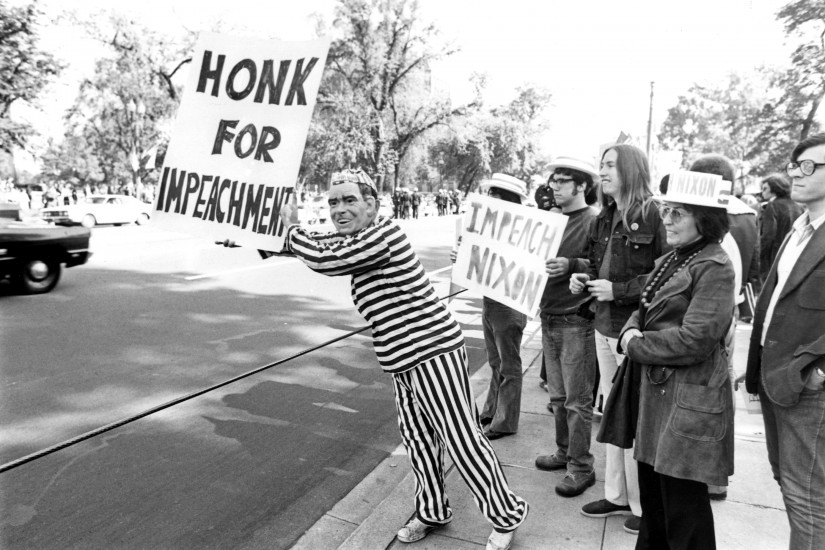 IA group of people holding signs stands on a sidewalk. A man in a striped prison suit and a Nixon mask leans out into the street, holding a "Honk for Impeachment" sign.