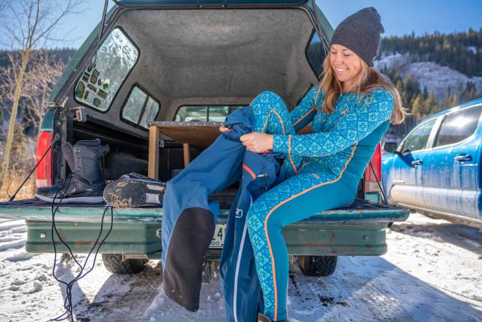 woman sitting on green truckbed in Kari Traa baselayers and beanie putting on pants and smiling