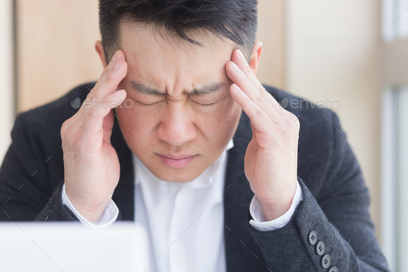 young asian man, office worker sitting at workplace holding hands massages forehead and head with