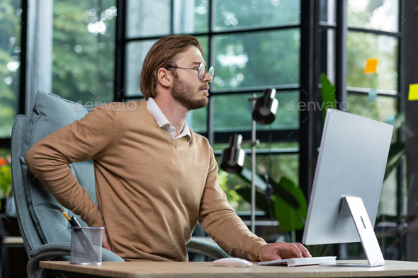 A young man is tired at the workplace in the office, massages his back with his hand, overtime work