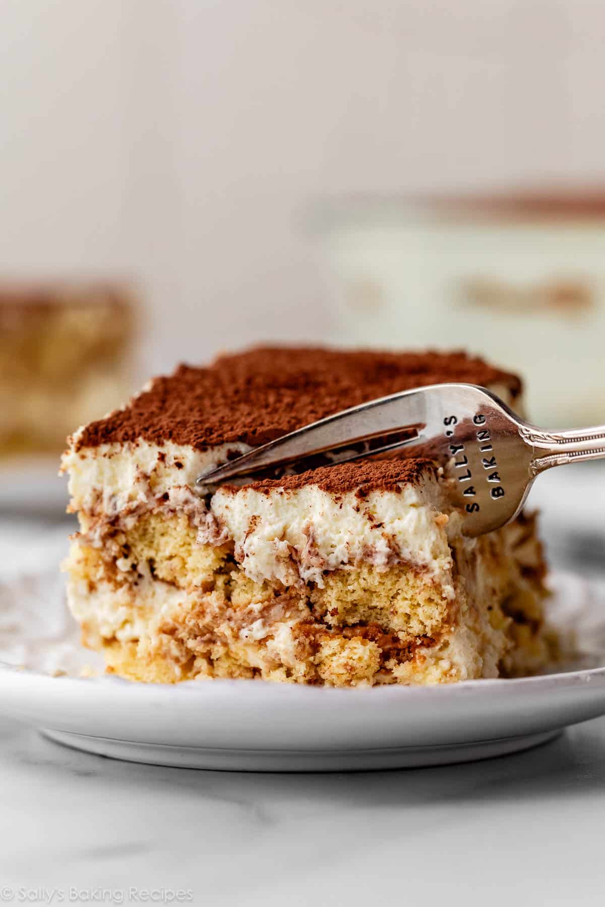 Slice of tiramisu on white plate with fork cutting through.