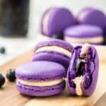 Blueberry macarons stacked on a wooden board with more macrons in the background.