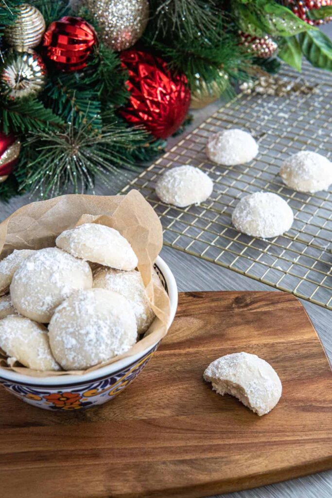 Snowball cookies in a bowl with one half eaten cookie on a wooden serving board. 