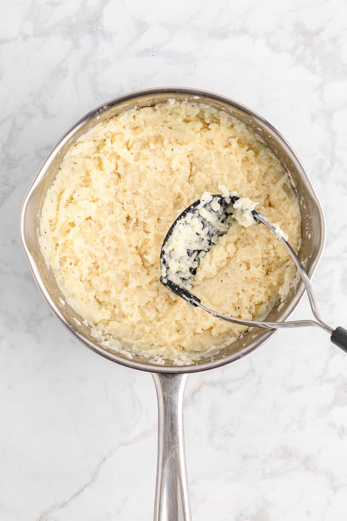 Mashing potatoes with a potato masher in a stainless steel saucepan.