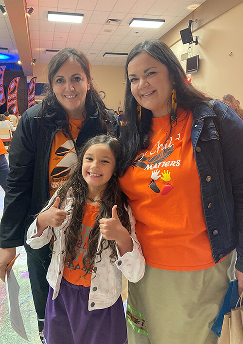 Charlene stands with her daughter and sister at an orange shirt day event. all are wearing orange shirts.