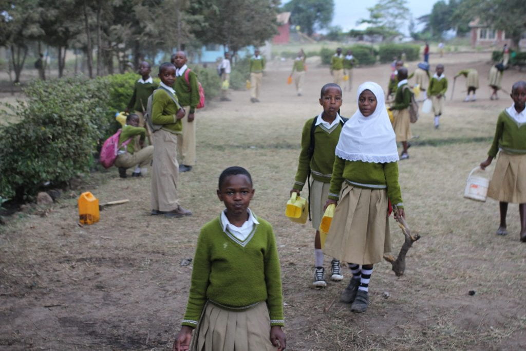 Several young orphans in matching uniforms stand in a grassy area, holding some types of tools. 