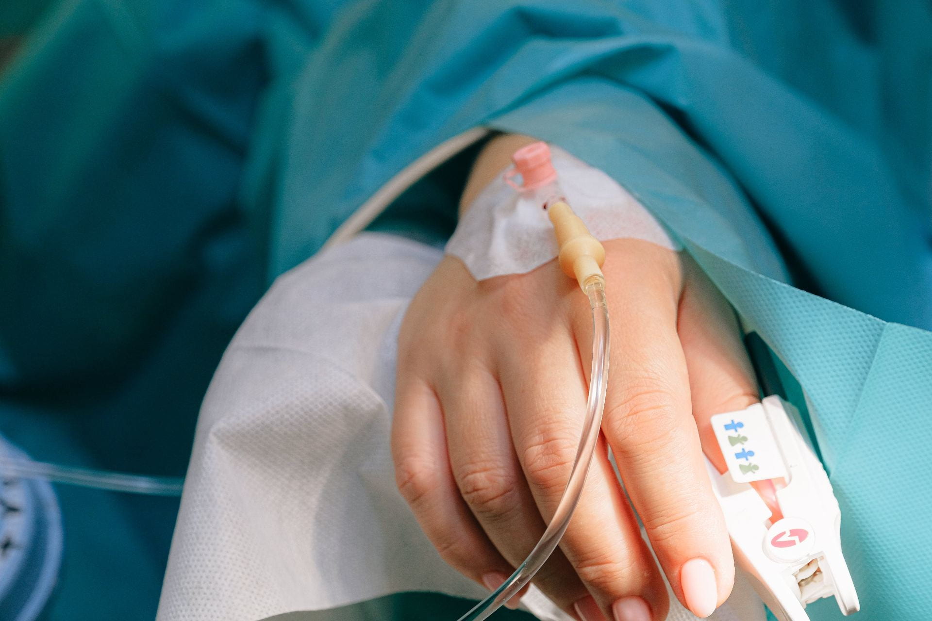 A hand with an IV line and a heart rate monitor on the thumb. The person’s arm is covered in a blue medical gown.