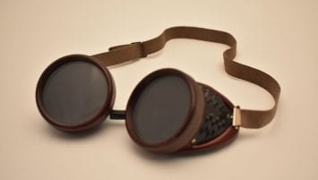 image of single pair of goggles. Dark green tinted lenses with a ridged outer rim; sides feature a metal mesh screen set into a Bakelite google frame. Adjustable elastic band connected to each side of the Bakelite frame.