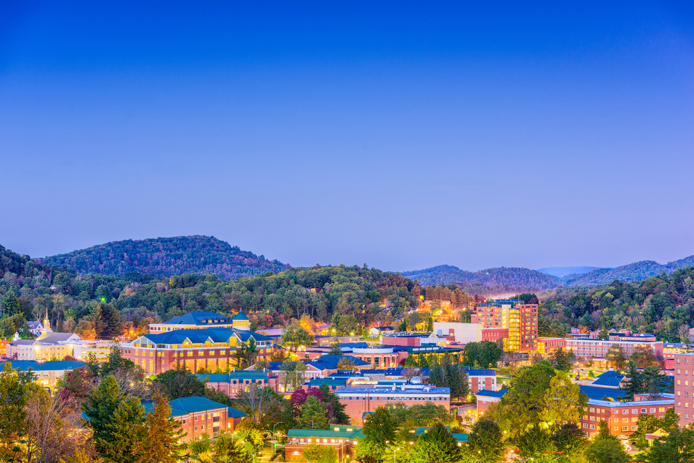 Buildings with lights glowing in the town of Boone, NC at dusk, surrounded by tree-filled mountains.