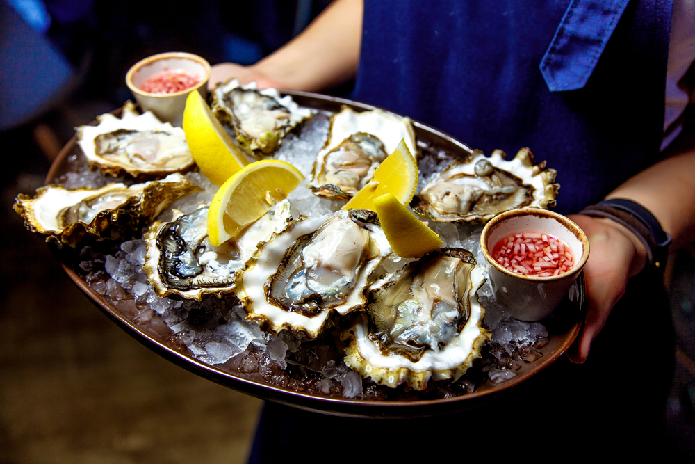 A person carrying a tray of oysters on ice. There are lemon wedges, and picked onions in small bowls as well. 