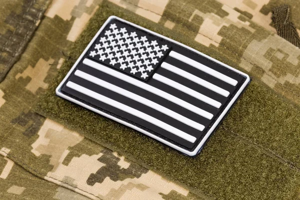 Vinyl US flag sticker in black and white color scheme with hook-and-loop fastener fixed on a military camouflage uniform, close-up in selective focus