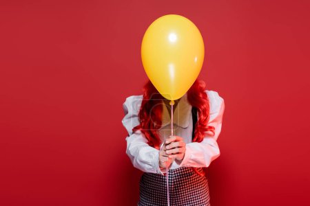 Foto de Mujer joven con el pelo de color que oscurece la cara con globo amarillo en la fiesta de halloween aislado en rojo - Imagen libre de derechos