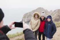 Hombre con cámara de teléfono fotografiando a la familia en ropa de abrigo en el acantilado con vistas al océano - foto de stock