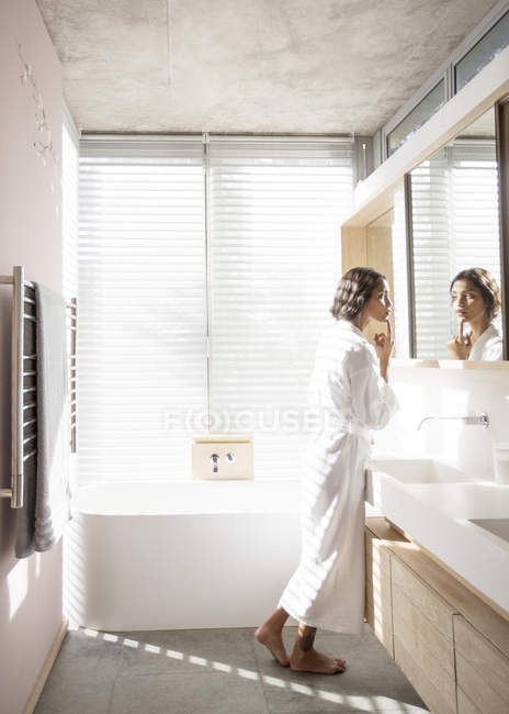 Woman in bathrobe examining face in bathroom mirror — Stock Photo