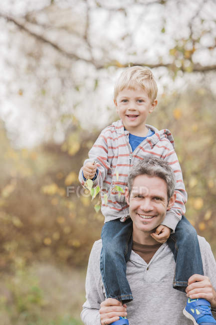 Portrait smiling father carrying toddler son on shoulders in autumn park — Stock Photo