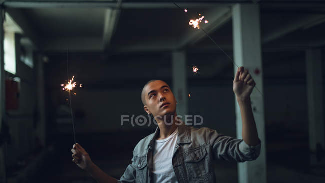 Vista frontale di un giovane ispano-americano che indossa una giacca grigia sopra una camicia bianca guardando lontano dalla fotocamera mentre tiene accese scintille su entrambe le mani all'interno di un magazzino vuoto — Foto stock