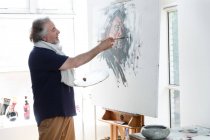 Side view of smiling mature artist painting self-portrait on easel in studio — Stock Photo