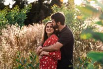 Pensive man hugging smiling pregnant wife on background of picturesque green park in sunny day — Stock Photo