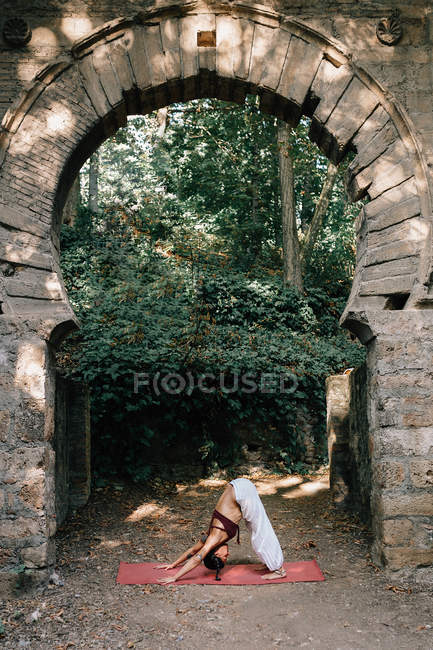 Woman standing in Downward-Facing Dog pose while doing yoga under old crumbling arch — Stock Photo