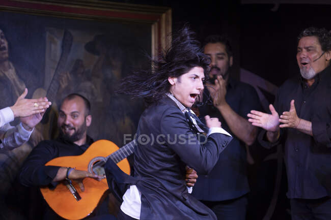 Uomo in costume nero danza flamenco vicino a musicisti maschi ispanici durante la performance contro la pittura sul palco scuro — Foto stock