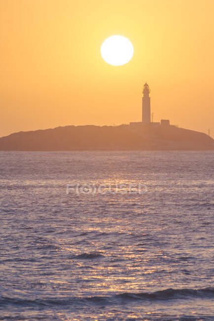 Paesaggio pittoresco di acqua increspata della costa di lavaggio dell'oceano con faro a molti piani situato a Faro de Trafalgar a Cadice in Spagna sotto il cielo arancione brillante all'alba — Foto stock