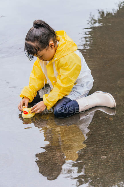 Affascinante bambino etnico in impermeabile che gioca con anatre di plastica che si riflettono nella pozzanghera increspata nel tempo piovoso — Foto stock