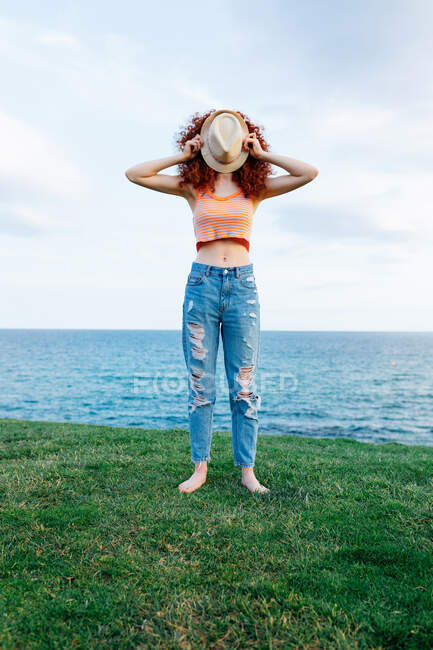 Corpo pieno di femmina scalza irriconoscibile con riccioli lunghi di zenzero che coprono la faccia di cappello su costa erbosa di mare azzurro — Foto stock