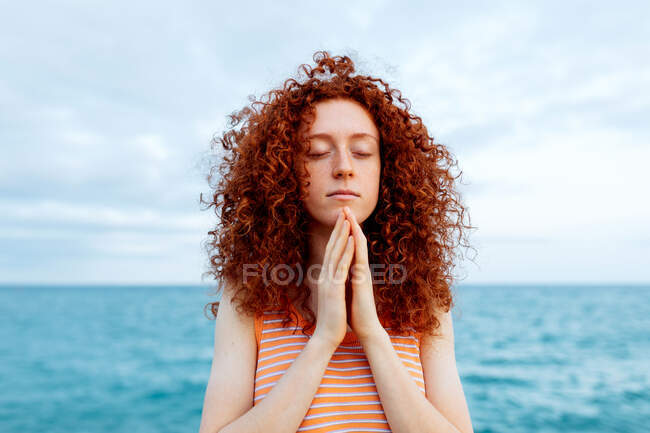 Donna pacifica in piedi con gli occhi chiusi sulla riva del mare e fare gesto namaste durante la meditazione — Foto stock