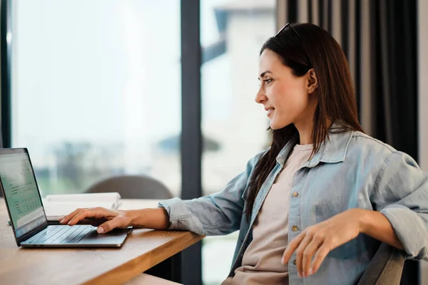 Attractive young smart woman working on laptop computer while spending time at home in the kitchen