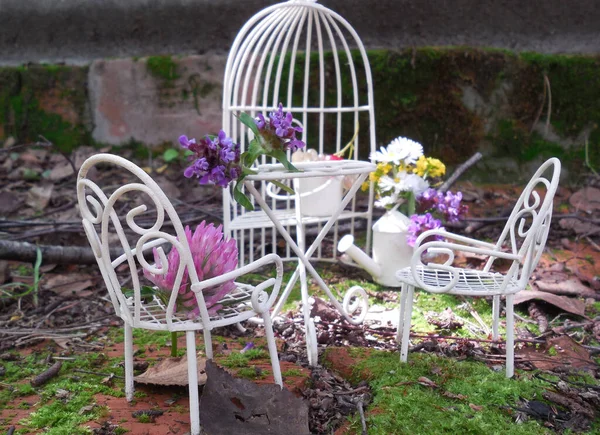 A miniature fairy garden on a platform of old bricks covered with moss and green leaves. Small white metal furniture: gazebo, table and chairs with backs. Flowers and berries.