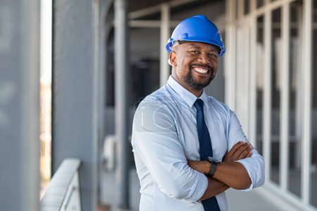 Foto de Retrato del arquitecto afroamericano en la obra con los brazos cruzados mirando a la cámara. Gerente de construcción de confianza en la ropa formal que lleva hardhat azul. Ingeniero civil maduro exitoso en el sitio de construcción con espacio de copia
. - Imagen libre de derechos