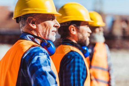 Foto de Constructores en chalecos reflectantes y cascos duros de pie juntos en el sitio de construcción, mirando a la distancia - Imagen libre de derechos