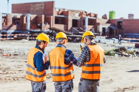 Foto de Tres trabajadores con sombreros y chalecos reflectantes de pie en la obra - Imagen libre de derechos