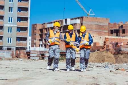 Foto de Tres trabajadores en chalecos reflectantes y sombreros caminando juntos a través del sitio de construcción y discutiendo planes de construcción - Imagen libre de derechos