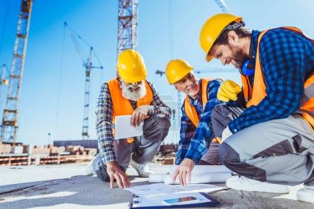 Foto de Tres trabajadores de la construcción sentados en concreto en el sitio de construcción, discutiendo planes de construcción - Imagen libre de derechos