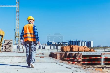 Foto de Constructor en chaleco reflectante y hardhat caminando a través de un sitio de construcción con rollos de planos en sus manos - Imagen libre de derechos