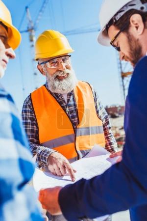 Foto de Trabajador de la construcción en uniforme protector discutiendo planes de construcción con hombre de negocios en hardhat y traje - Imagen libre de derechos