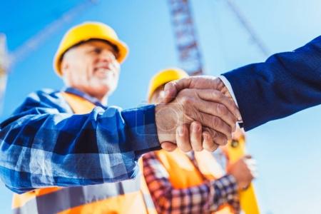 Foto de Trabajador de la construcción en uniforme de protección estrechando la mano con el hombre de negocios en la obra - Imagen libre de derechos