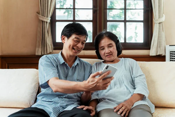 Happy older people sitting on a couch using smartphone. Grandmother listen to music with wireless headphone controlled by smartphone. Selfie.