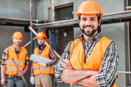 Foto de Apuesto constructor sonriente de pie en el sitio de construcción con los brazos cruzados mientras sus colegas de pie borrosa en el fondo - Imagen libre de derechos