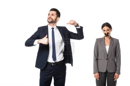 Foto de Arrogante hombre de negocios en traje apuntando con los pulgares a sí mismo cerca de la mujer con cinta adhesiva en la boca aislado en blanco - Imagen libre de derechos