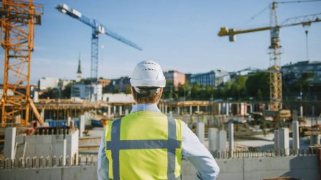 Foto de El Confident Bearded Head Civil Engineer-Architect in Sunglasses está parado afuera con su espalda a la cámara en un sitio de construcción en un día brillante. El hombre lleva un sombrero duro, una camisa y un chaleco de seguridad. - Imagen libre de derechos