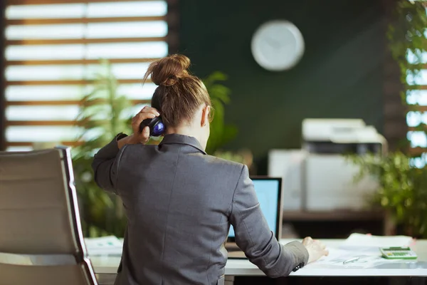 Sustainable workplace. Seen from behind modern small business owner woman in a grey business suit in modern green office using massage roller on neck.