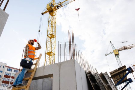 Foto de Hombre trabajador de mantenimiento con casco de seguridad y chaleco naranja escalando escalera de madera y sosteniendo la caja de herramientas en el sitio de construcción - Imagen libre de derechos