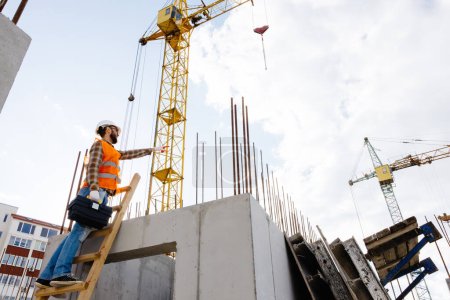 Foto de Hombre trabajador de mantenimiento con casco de seguridad y chaleco naranja escalando escalera de madera y sosteniendo la caja de herramientas en el sitio de construcción - Imagen libre de derechos